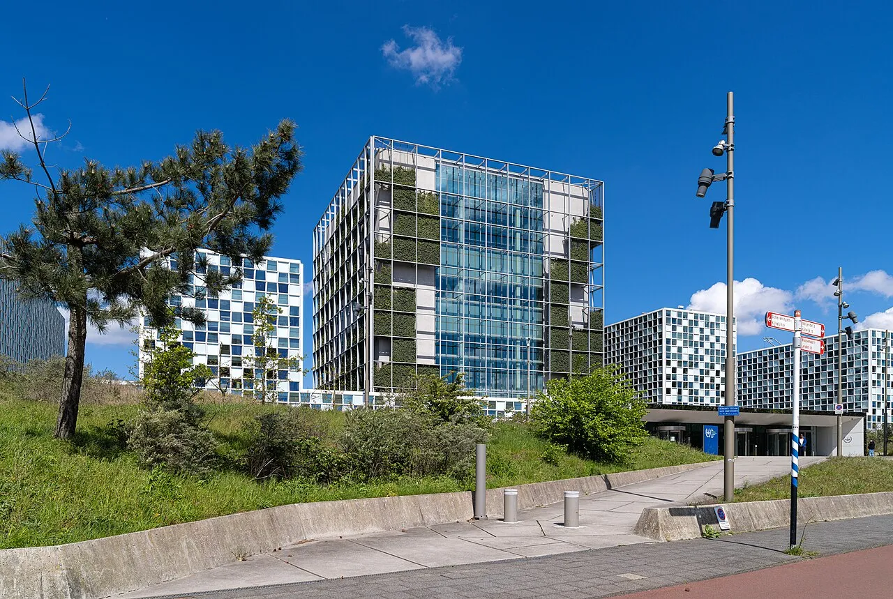 Het gebouw van het Internationaal Strafhof in Den Haag, een glazen toren met groene gevelbeplanting naast witte blokken, onder een blauwe lucht.